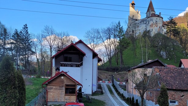 Un pueblo rural con una iglesia destacada en una colina, una casa de madera con balcón y un coche rojo estacionado al frente.