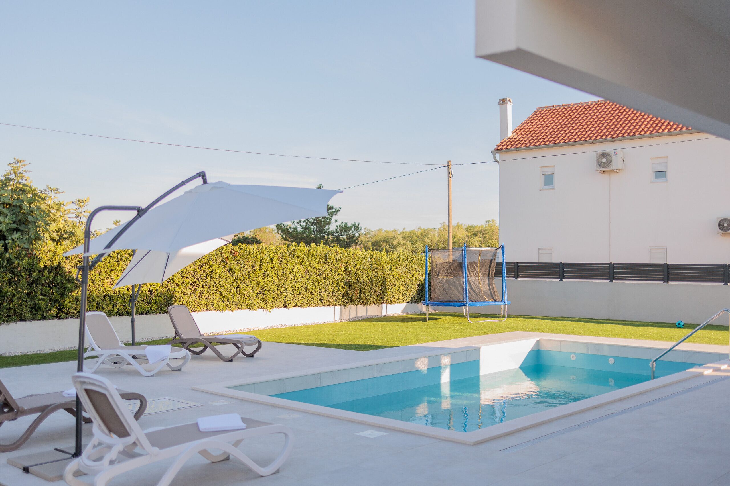 A swimming pool with lounge chairs and umbrellas, surrounded by a hedge and a white building with a red roof.