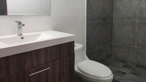 Un bany amb un lavabo blanc, un moble de tocador de fusta fosca, un vàter i una dutxa amb finestra.