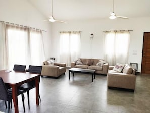 A living room with a sofa set, a coffee table, and a dining table with chairs. There are two ceiling fans and a door visible.