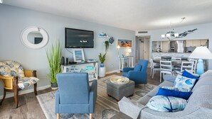 A living room with a TV, blue armchairs, a sofa, and a dining area with a table and chairs. - S Hampton 1403 1 Br Oceanview in Kingston, Pool (Myrtle Beach)