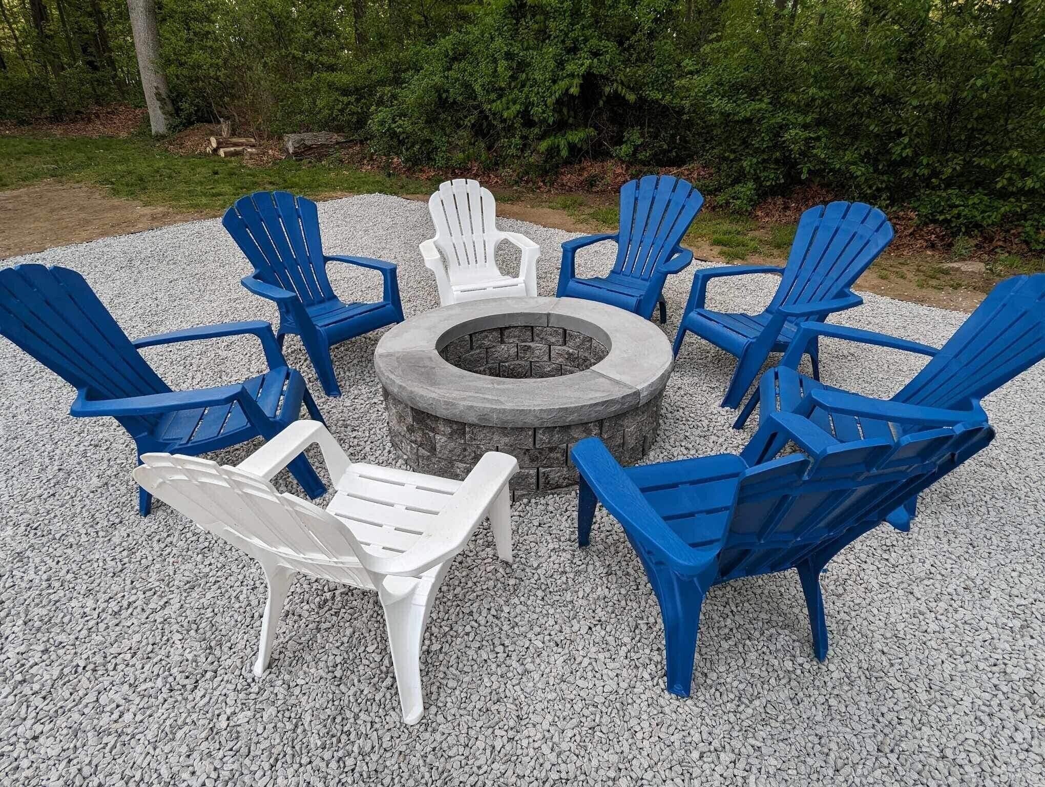 Disposición circular de asientos con un hogar central, rodeado de sillas Adirondack azules y blancas sobre una superficie de grava, ubicado en un área boscosa.