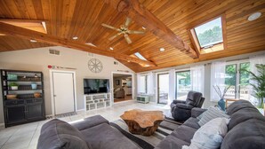 Een ruime woonkamer met een houten plafond, een grote televisie en een gezellige zithoek met een bank en fauteuils.