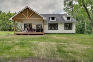 Uma casa moderna com varanda coberta e um deck espaçoso, cercada por um gramado e árvores.