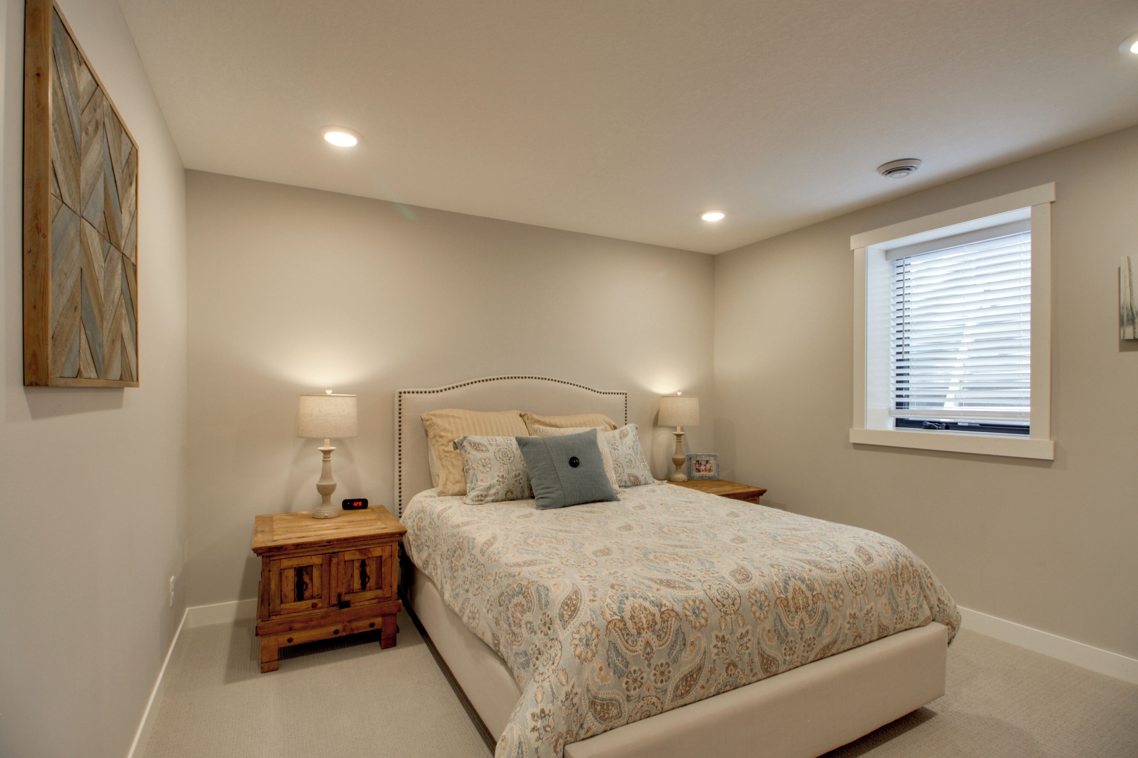 A bedroom with a bed, two bedside tables, a nightstand, a window with blinds, and a wall-mounted artwork.
