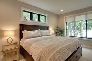 Quarto com uma cama grande, mesa de cabeceira com abajur, janela com persianas e vista para árvores.