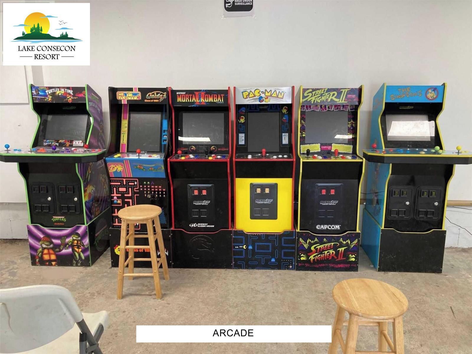 Een rij spelautomaten met verschillende spellen, waaronder 'Turtles in Time', 'Midway Mortal Kombat', 'Pac-Man', 'Street Fighter II' en 'The Simpsons'. Er staan twee krukken voor de spelautomaten.