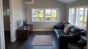 A living room with a black leather couch, a wooden desk with a TV, a large window with a view of a house and beach, and a ceiling-mounted air conditioner. - LAKEFRONT COTTAGE #5 - PRINCE EDWARD COUNTY - LAKE CONSECON RESORT (Prince Edward)