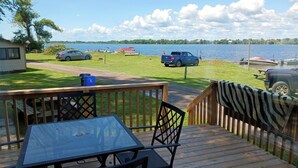 A wooden deck with a table and chairs, overlooking a lake with boats and a grassy area with parked cars. - LAKEFRONT COTTAGE #5 - PRINCE EDWARD COUNTY - LAKE CONSECON RESORT (Prince Edward)