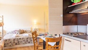 Una habitación pequeña con cama, mesa, sillas, lámpara y kitchenette.