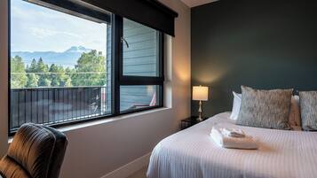A hotel room with a bed, a chair, a nightstand with a lamp, and a window with a view of mountains and trees.
