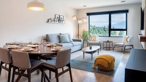 A modern living room with a dining area, a sofa, a coffee table, and a large window with a view. - Northern Exposure by Revelstoke Vacations (Revelstoke)