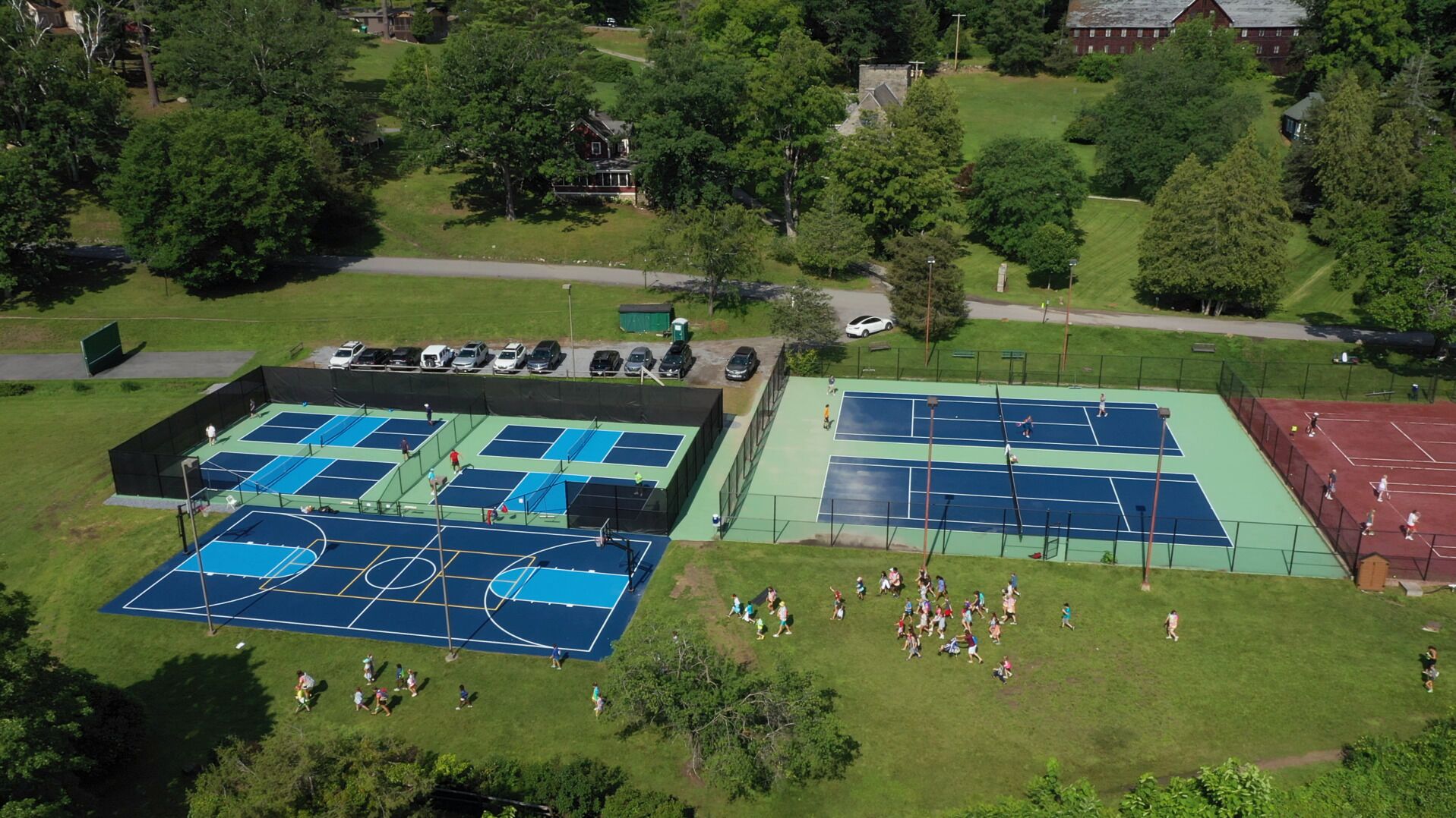 An aerial view of a sports complex with multiple tennis courts, surrounded by trees and residential buildings.