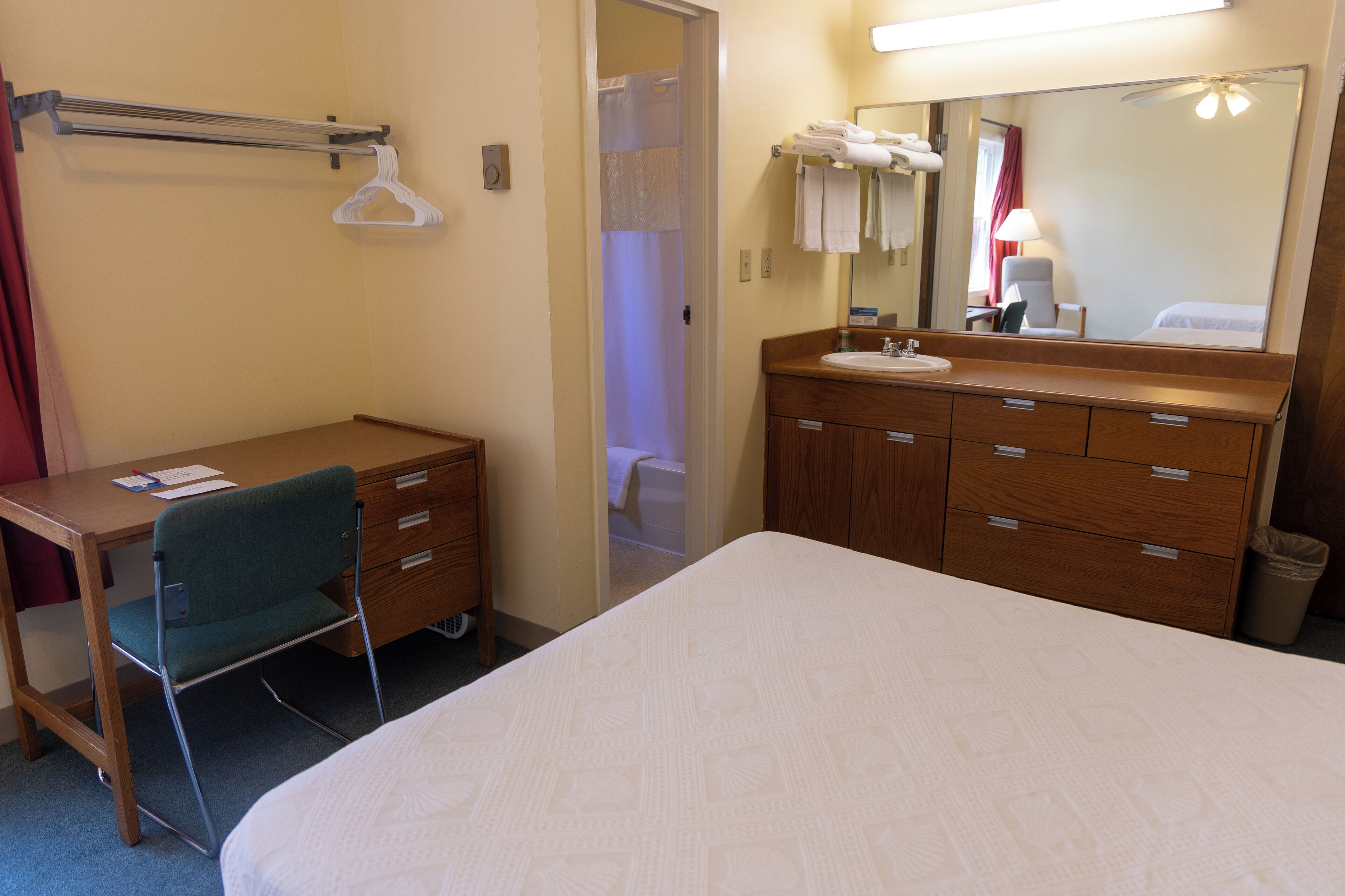 Une chambre d’hôtel comprenant un lit, un bureau avec une chaise, une commode en bois avec des tiroirs, un miroir et une salle de bain visible à travers une porte ouverte.