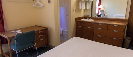 A hotel room with a bed, a desk with a chair, a wooden dresser with drawers, a mirror, and a bathroom visible through an open door.