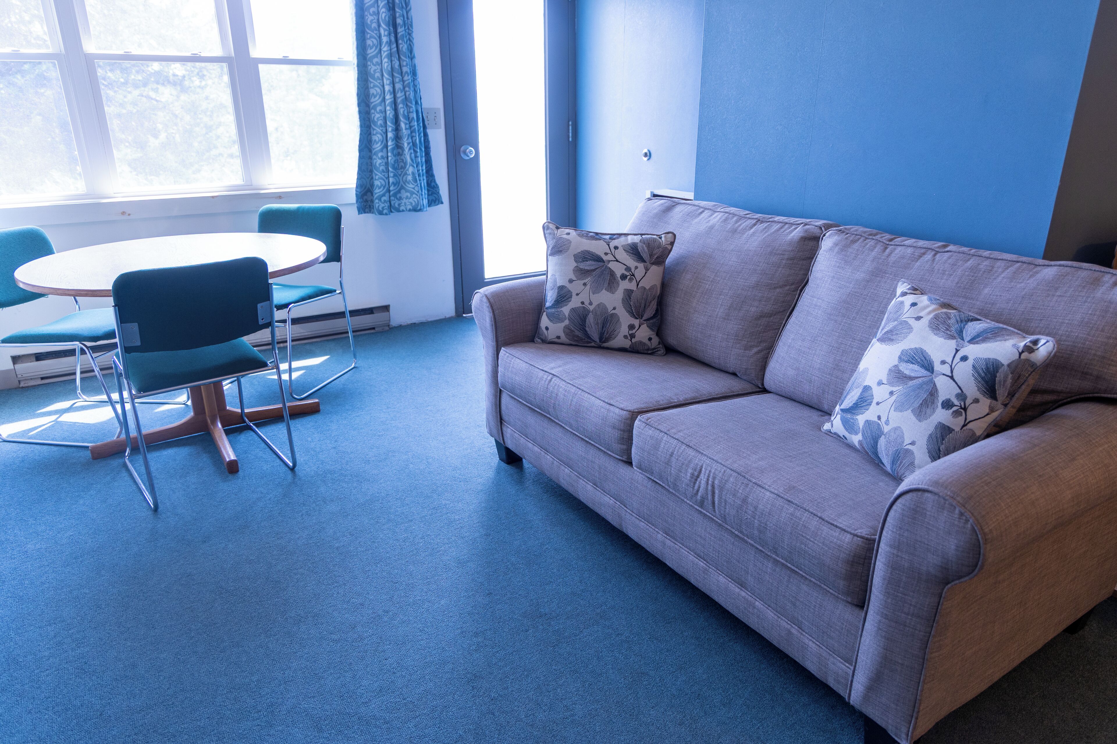 A room with a grey sofa, two matching cushions, a round table with two chairs, and a large window with blue curtains.