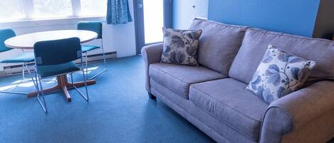 A room with a grey sofa, two matching cushions, a round table with two chairs, and a large window with blue curtains.