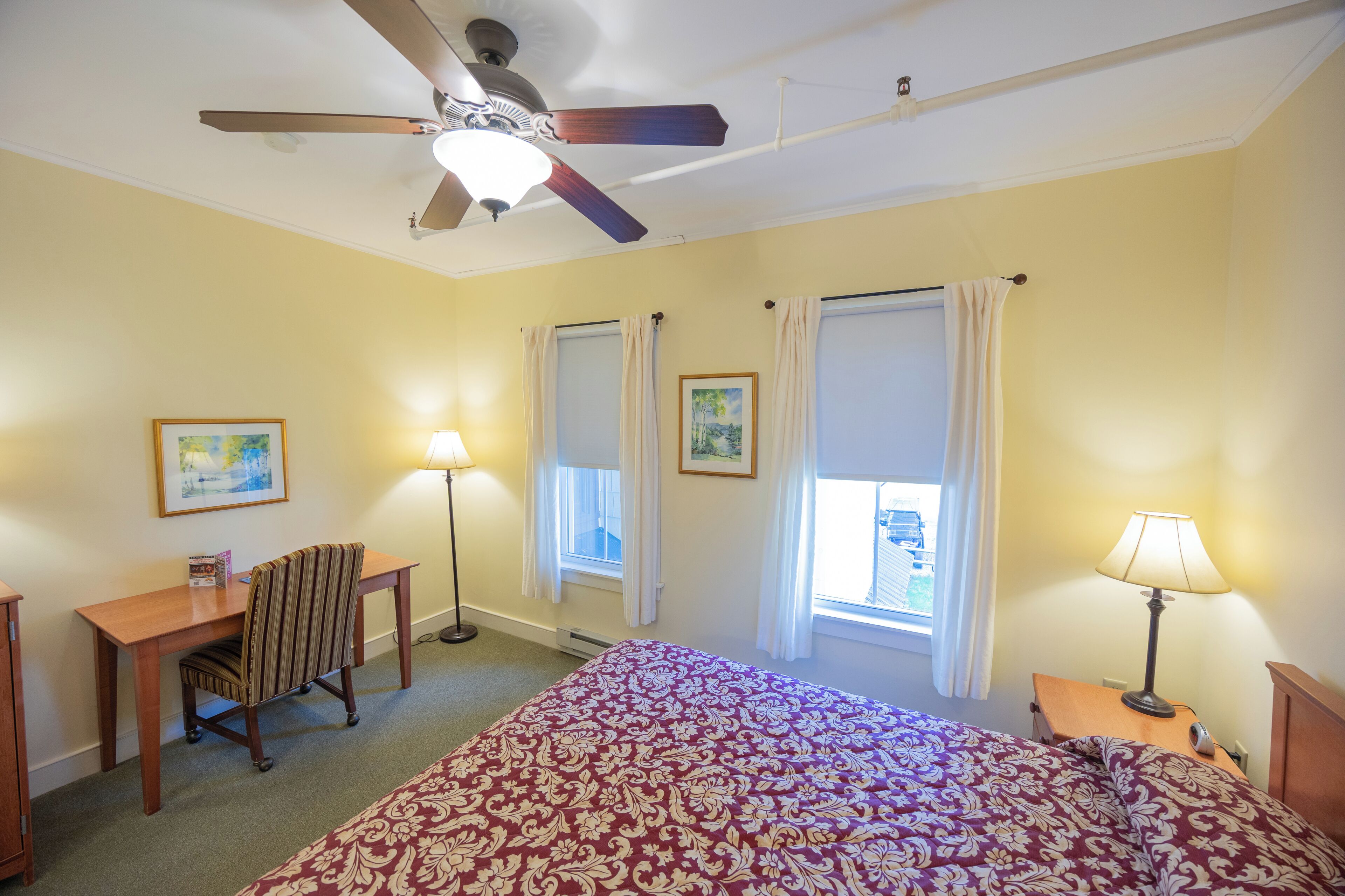Habitación de hotel con cama, escritorio, silla, ventilador de techo, dos ventanas con cortinas y dos lámparas de pared.