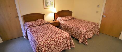 A room with two beds, each with a wooden headboard, a nightstand with a lamp, and a framed picture on the wall.