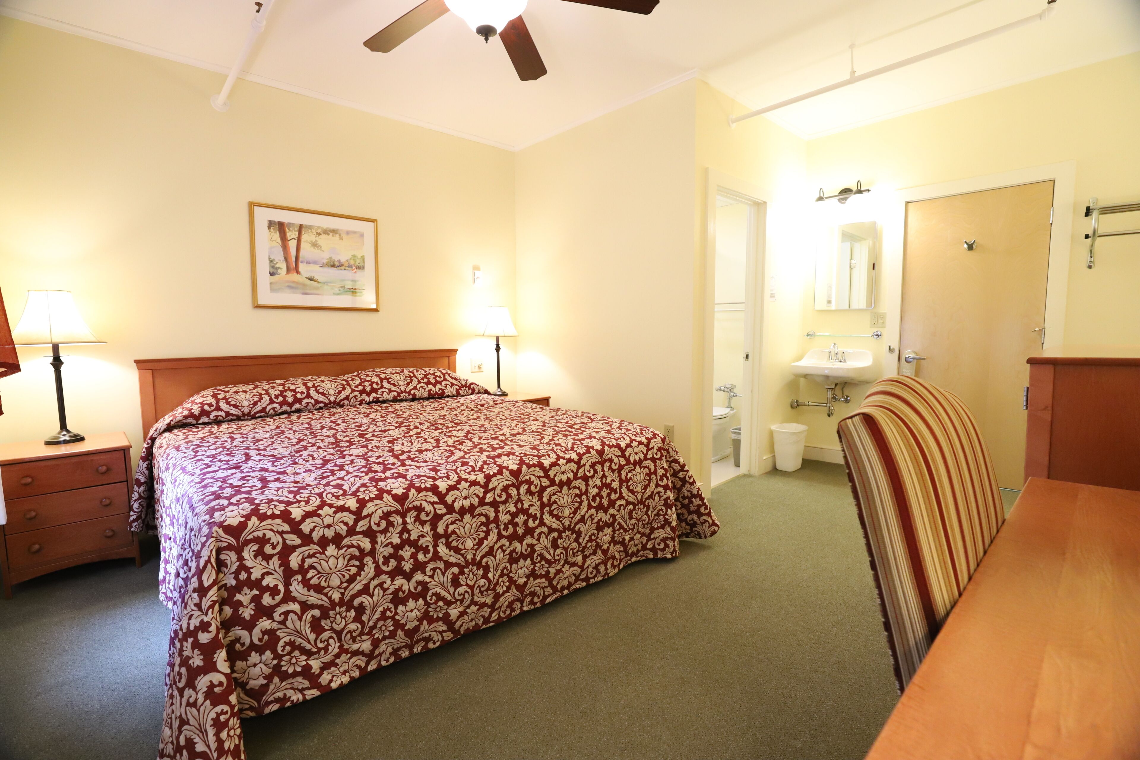 Une chambre à coucher avec un lit, des lampes de chevet, un ventilateur de plafond, un tableau, une salle de bain avec un lavabo et un miroir, et une table en bois.