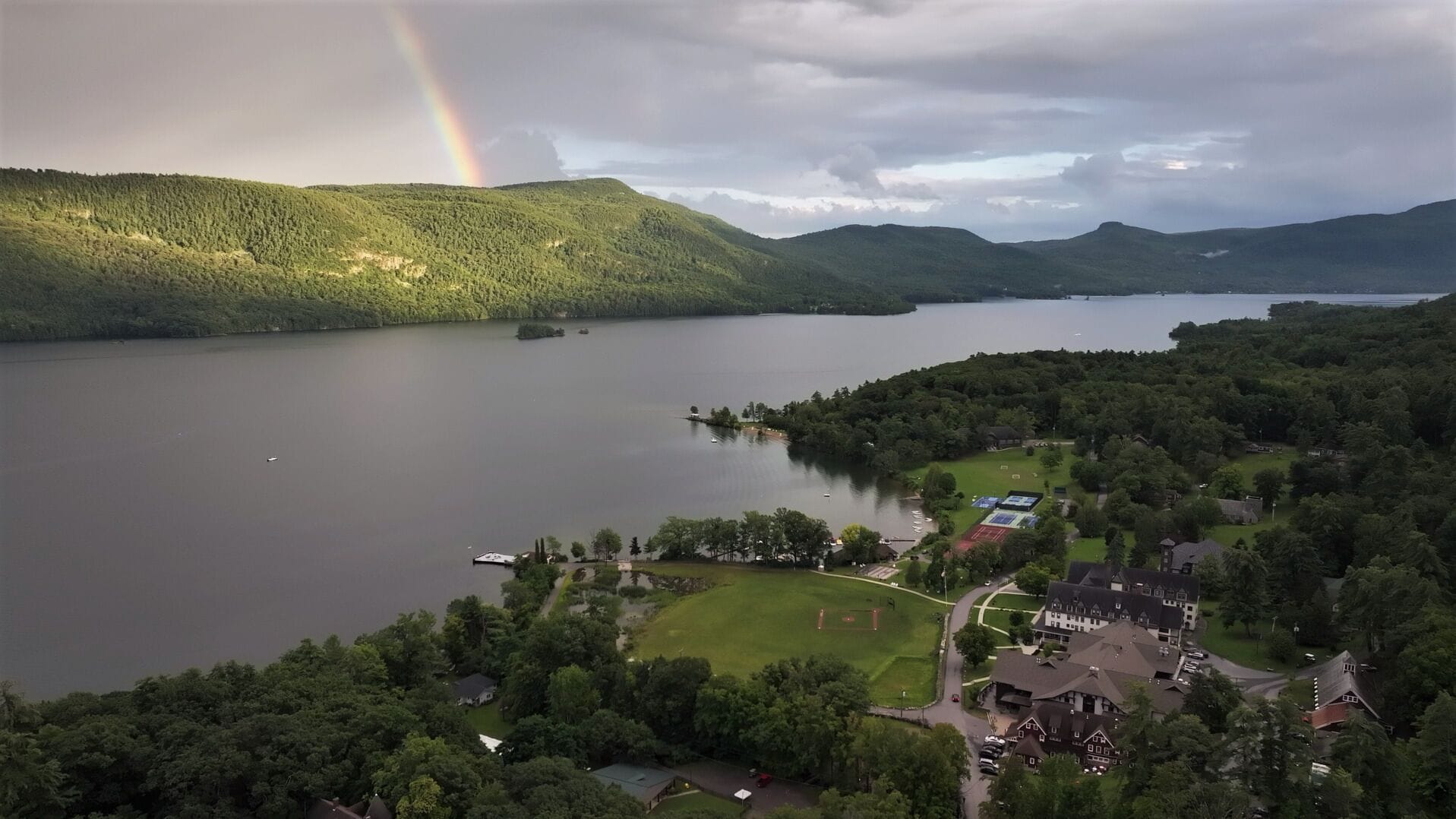 A lake surrounded by forested hills with a rainbow, a small dock, and a building with a large lawn.