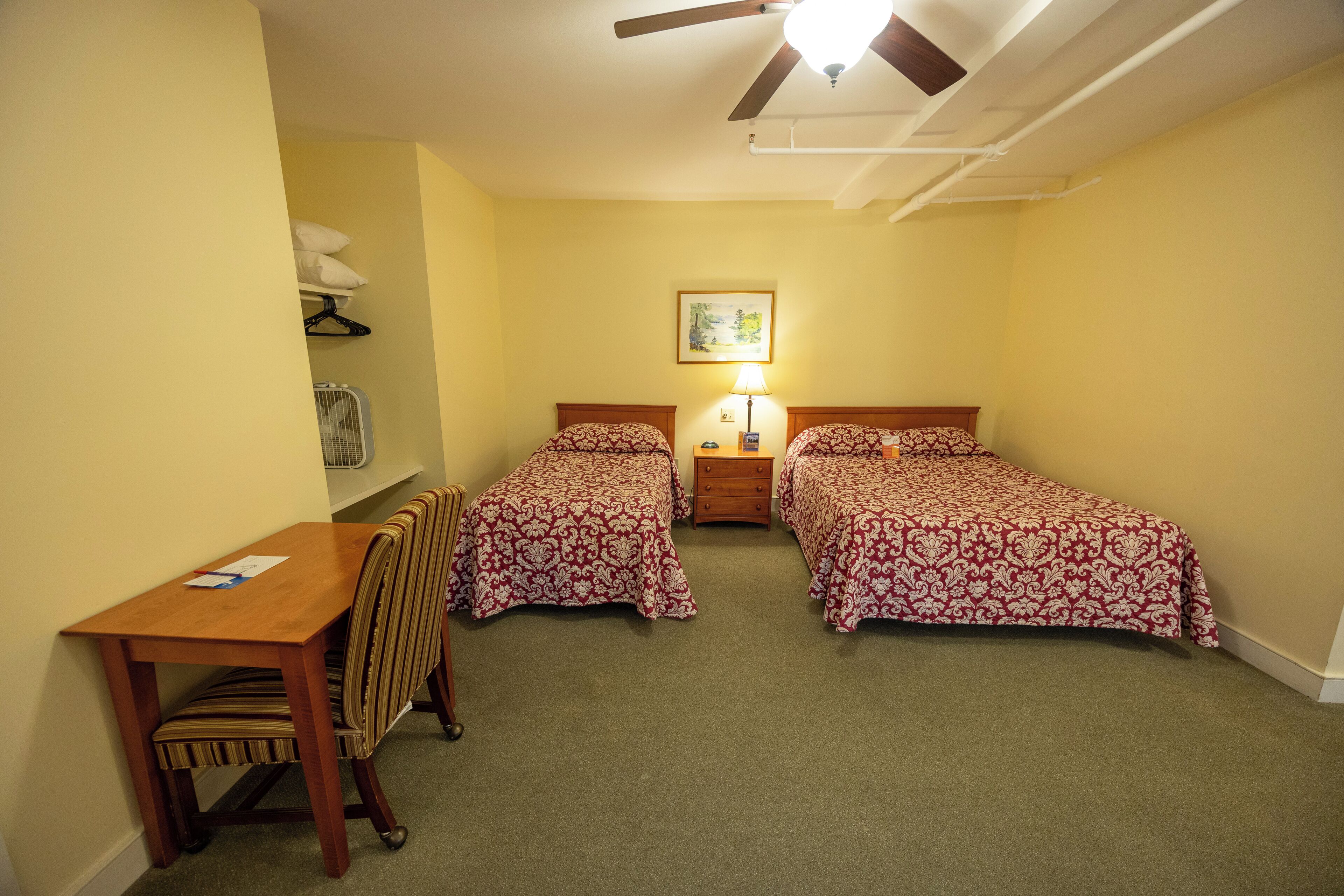 A room with two beds, a desk, a chair, a ceiling fan, a lamp, and a painting on the wall.