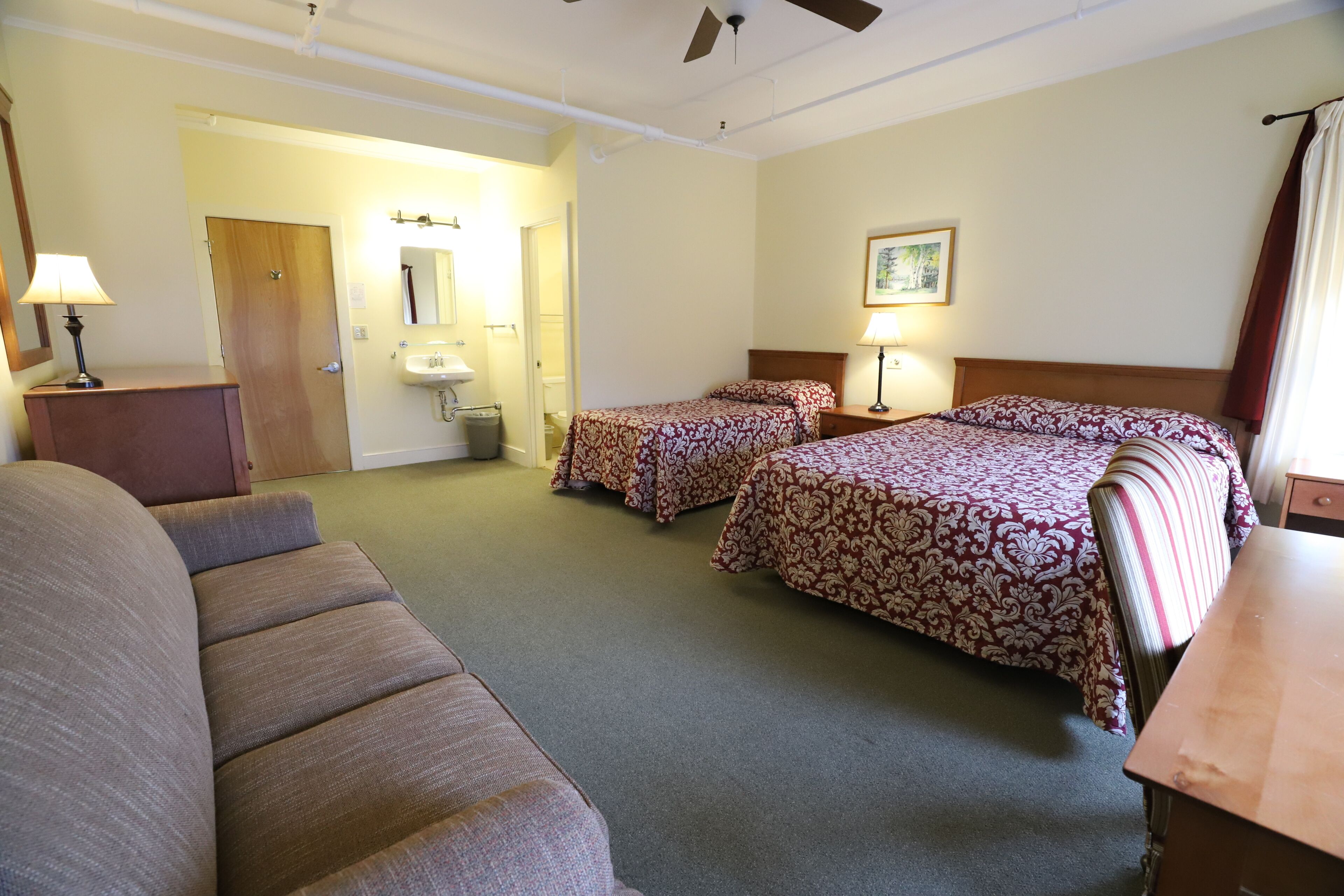 A hotel room with a sofa, two single beds, a wooden headboard, a bedside table with a lamp, a mirror, and a bathroom with a sink and toilet.