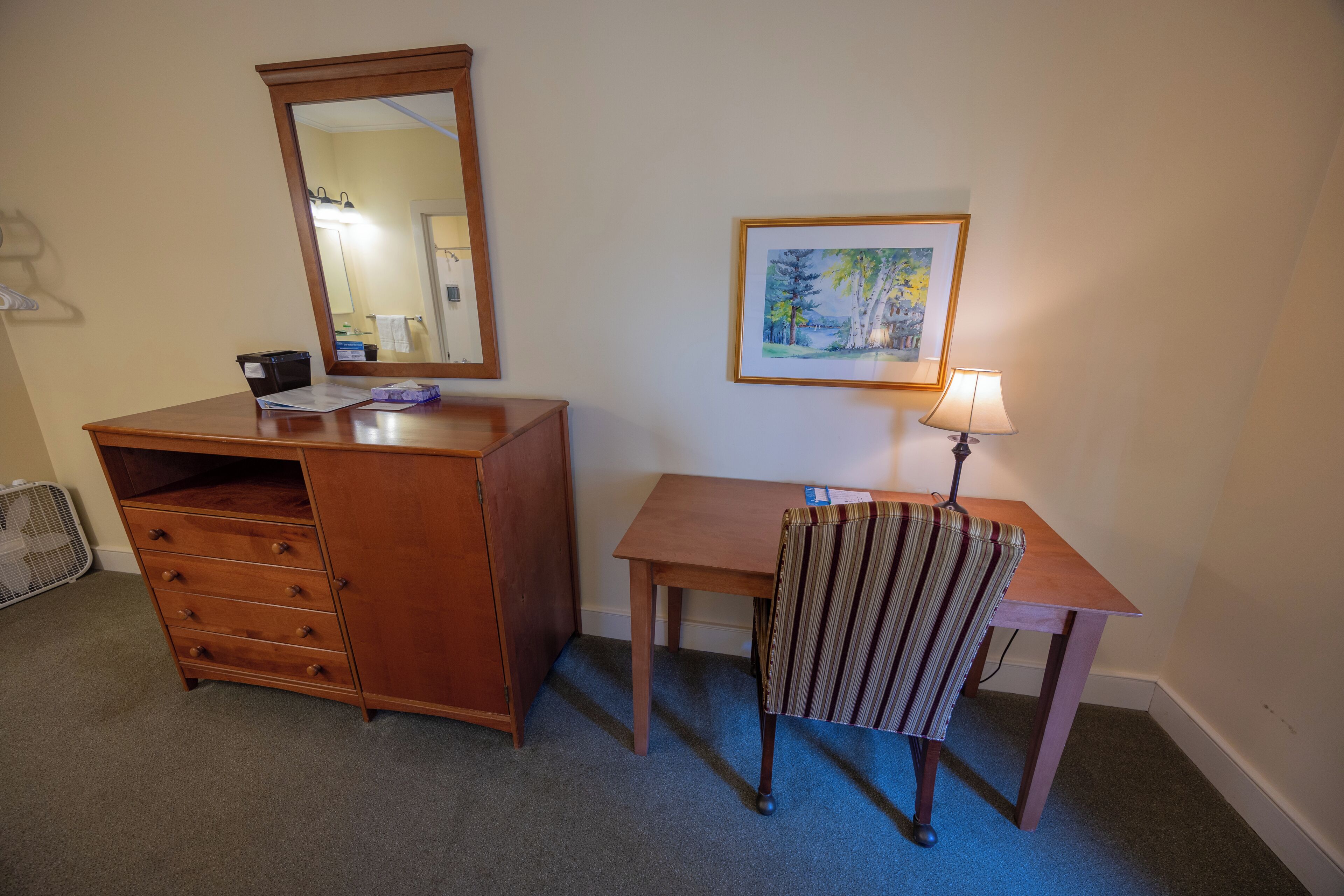 Una habitación con un escritorio de madera, una silla, un aparador con cajones, un espejo, un cuadro en la pared y una lámpara.