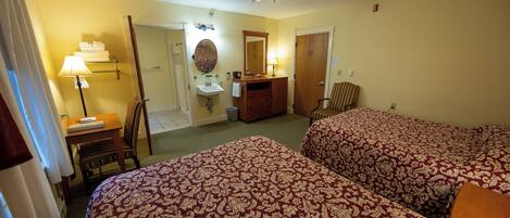 A hotel room with two beds, a desk with a lamp, a vanity with a mirror, and a ceiling fan.