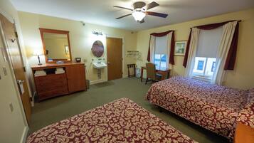 Une chambre d’hôtel avec deux lits, un ventilateur de plafond, un bureau, une chaise, une commode, un miroir et une fenêtre avec des rideaux.