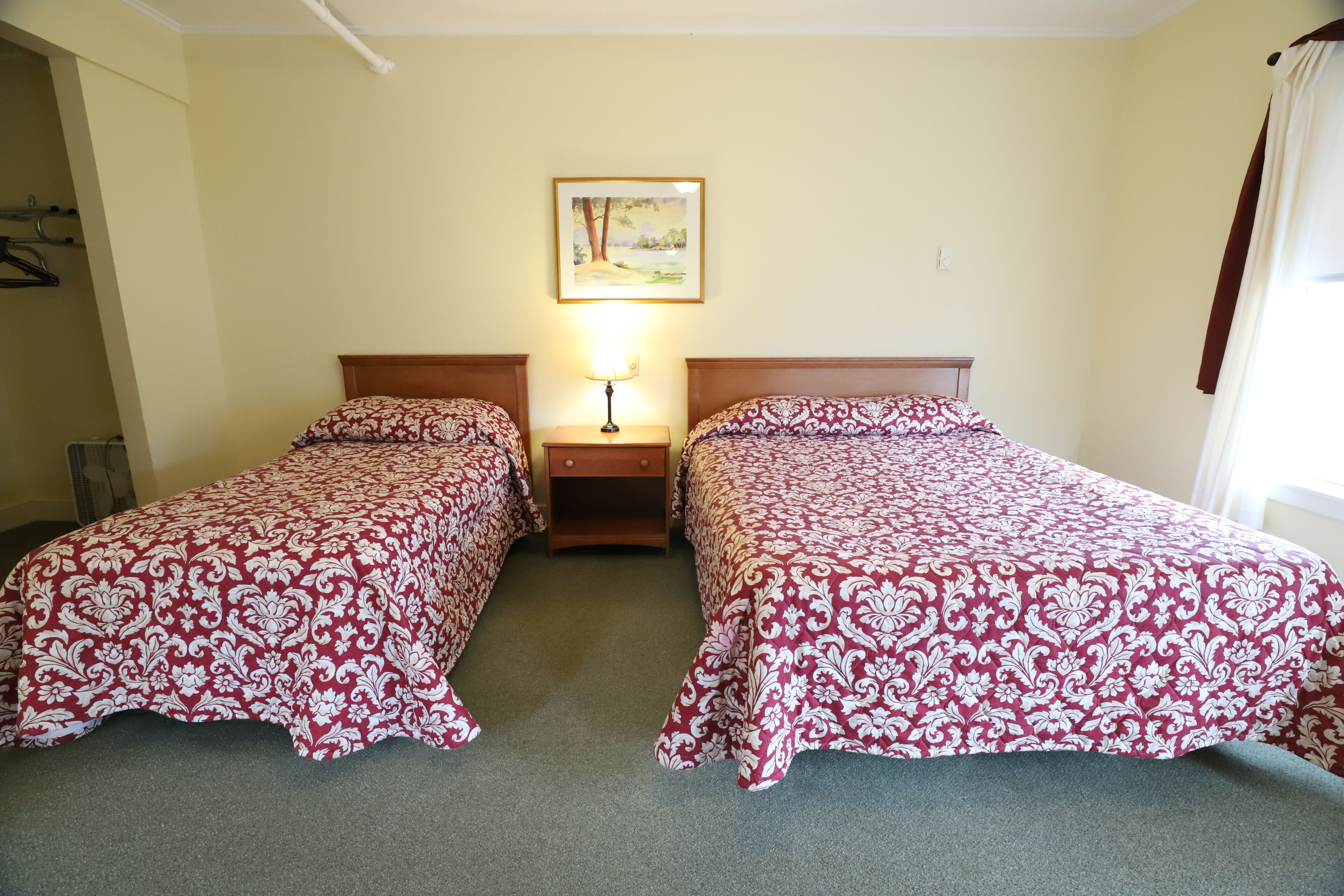 A hotel room with two single beds, a nightstand with a lamp, a painting on the wall, and a window with curtains.