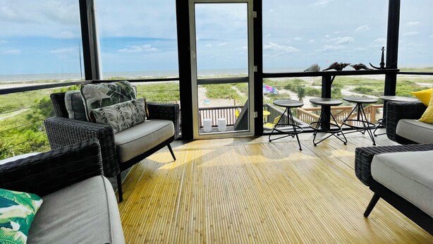 A covered outdoor seating area with wicker furniture, a view of the beach, and a wooden deck.