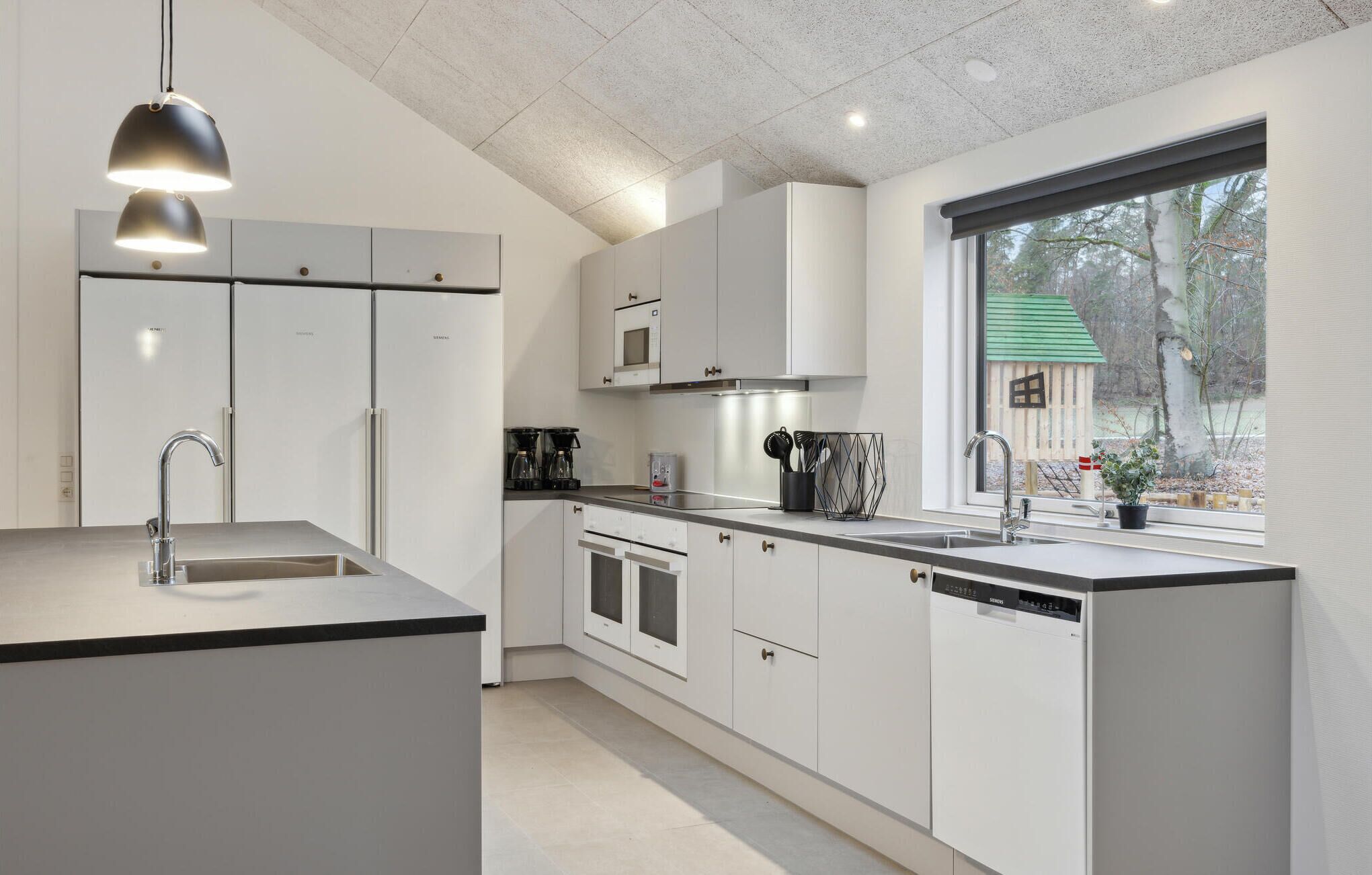 A modern kitchen with white cabinetry, a built-in oven, a dishwasher, and a window with a view of trees.