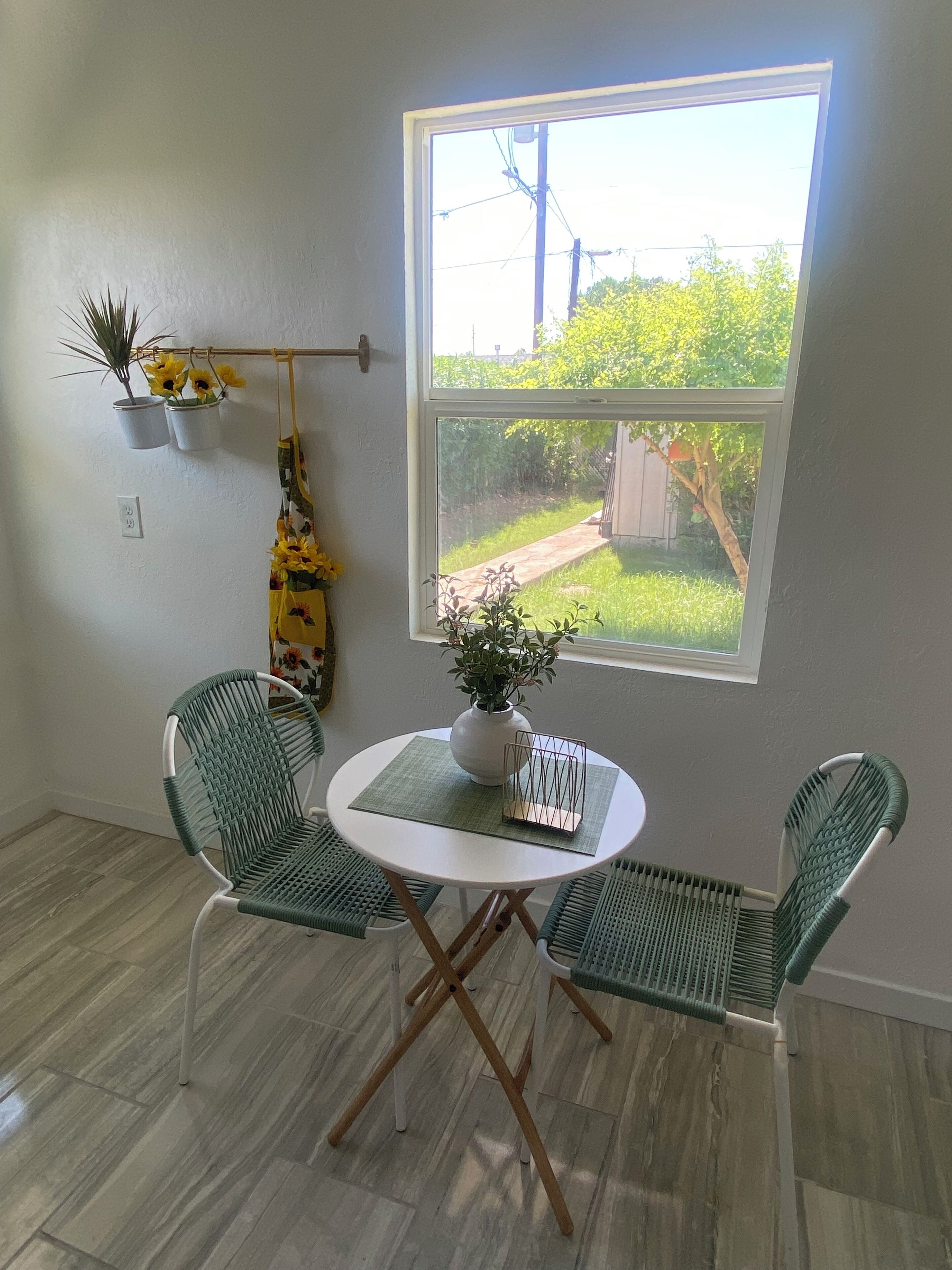 A small room with a round table and two chairs, a window with a view of a garden, and a wall-mounted shelf with plants.