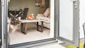 An open sliding glass door leading to a dining area with a table set for two, a bowl of fruit, and a sofa in the background.