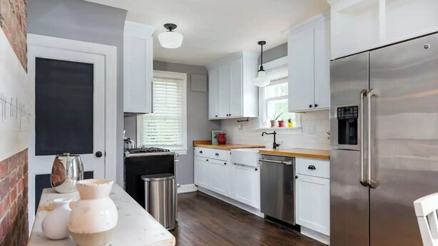 A modern kitchen with white cabinets, stainless steel appliances, a wooden countertop, and a brick wall.