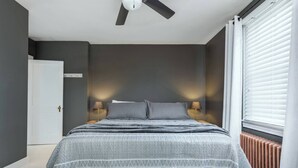 A bedroom with a bed, two bedside lamps, a ceiling fan, a radiator, and a window with blinds.