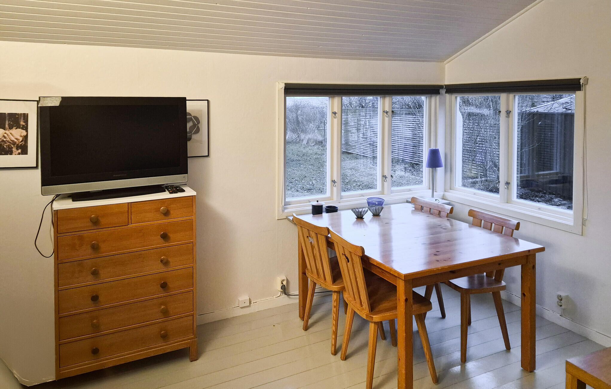A dining area with a wooden table and chairs, a television on a cabinet, and large windows with a view of trees.