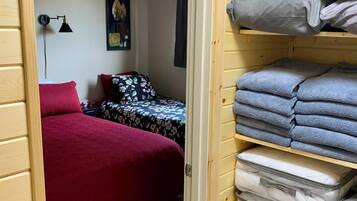 Une chambre avec un lit rouge, un ventilateur de plafond, une lampe fixée au mur et une étagère où sont pliées des serviettes et rangées des boîtes de rangement.