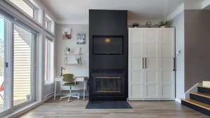 Een moderne woonkamer met een open haard, een televisie erboven, een bureau met stoel en een trap naar een hoger niveau.