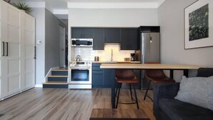 Een moderne keuken met donkere kasten, een roestvrijstalen oven en een magnetron. Er is een houten eettafel met twee barstoelen. Een grijze bank is op de voorgrond zichtbaar.