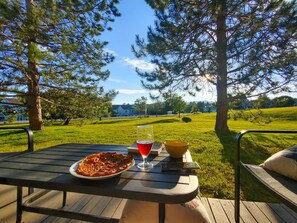 Spisebord udendørs med pizza, et glas rødvin og en skål på en terrasse med græsplæne og træer i baggrunden.