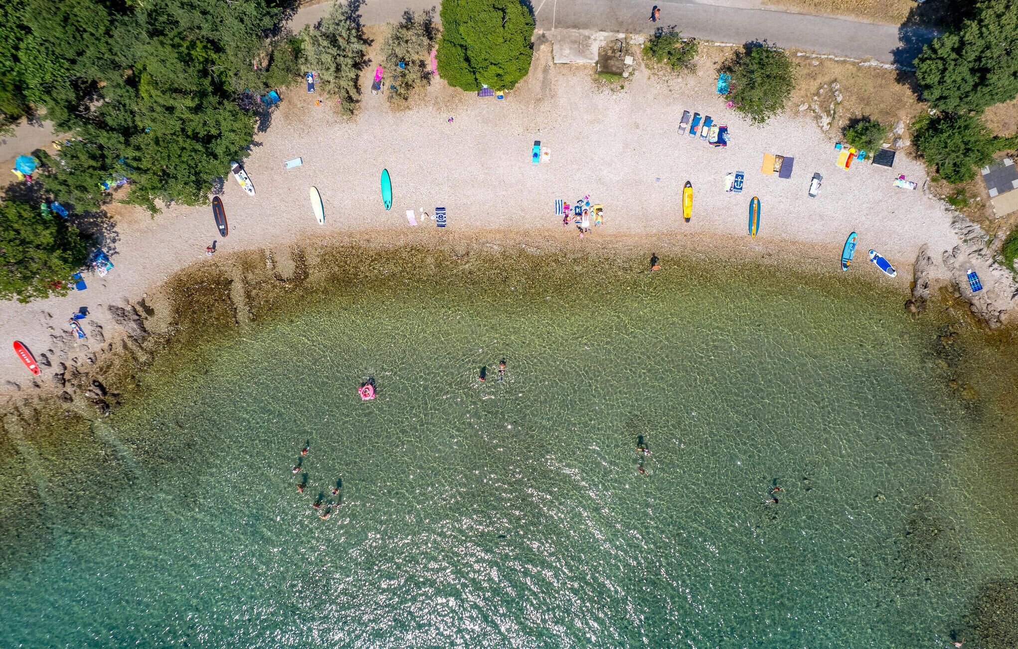 Luchtbeeld van een strand met helder water, mensen die zwemmen en diverse activiteiten op het strand.