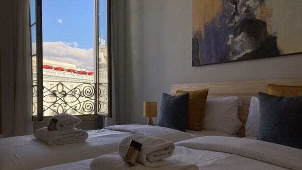 A hotel room with two beds, a large window with a view of buildings, and a painting on the wall. - Housing Sol by Sharing Co (Madrid)