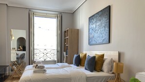 A modern bedroom with a large bed, a bedside table with a lamp, a wardrobe, and a view of the city through a window. - Housing Sol by Sharing Co (Madrid)