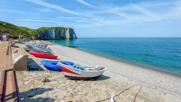 Ein Kiesstrand mit angelegten Booten, eine Klippe mit einer Höhlenöffnung und ein klares, blaues Meer.