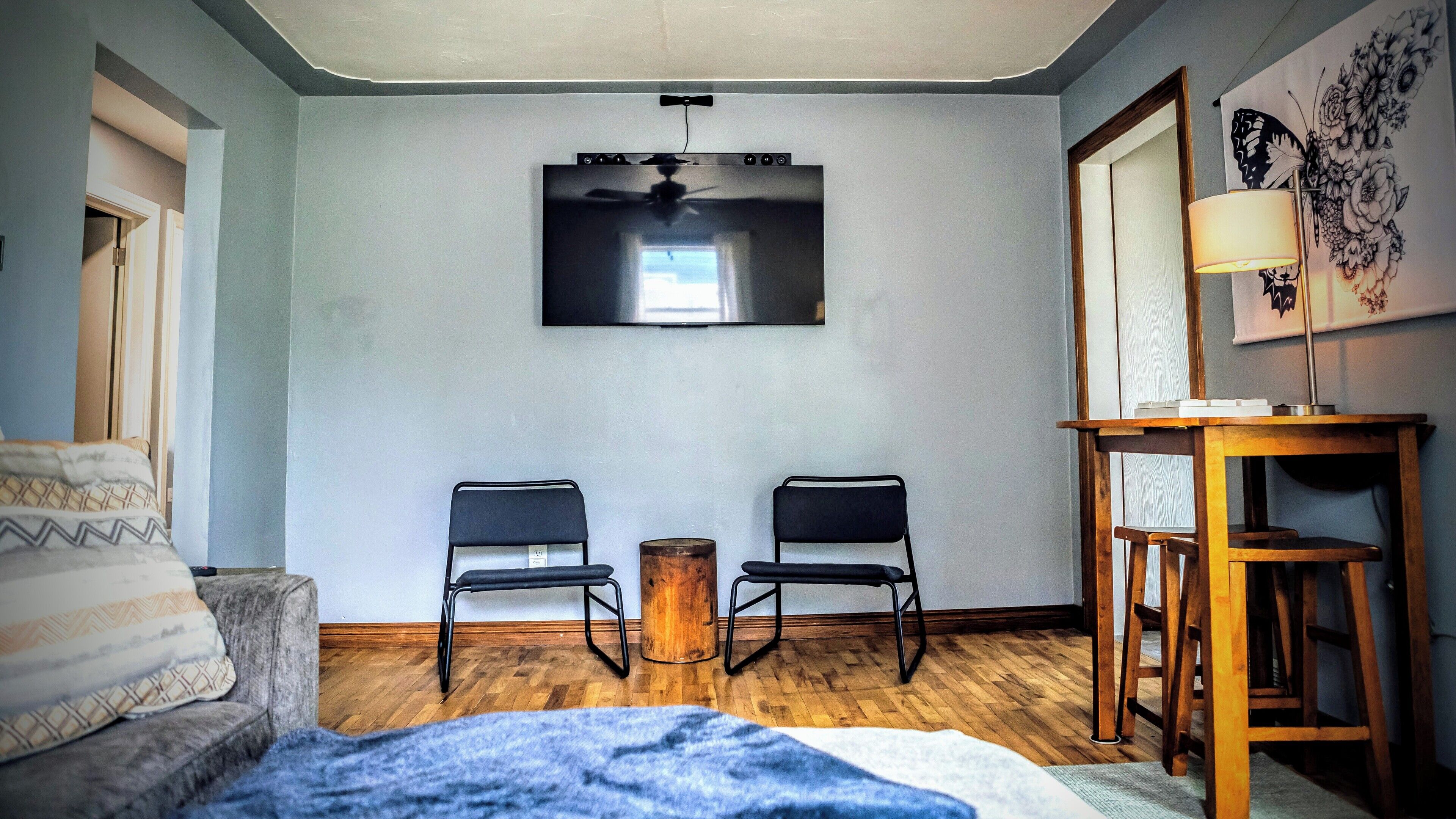 Et værelse med et fladskærms-tv på væggen, to sorte stole, et træ sidebord og en lampe.