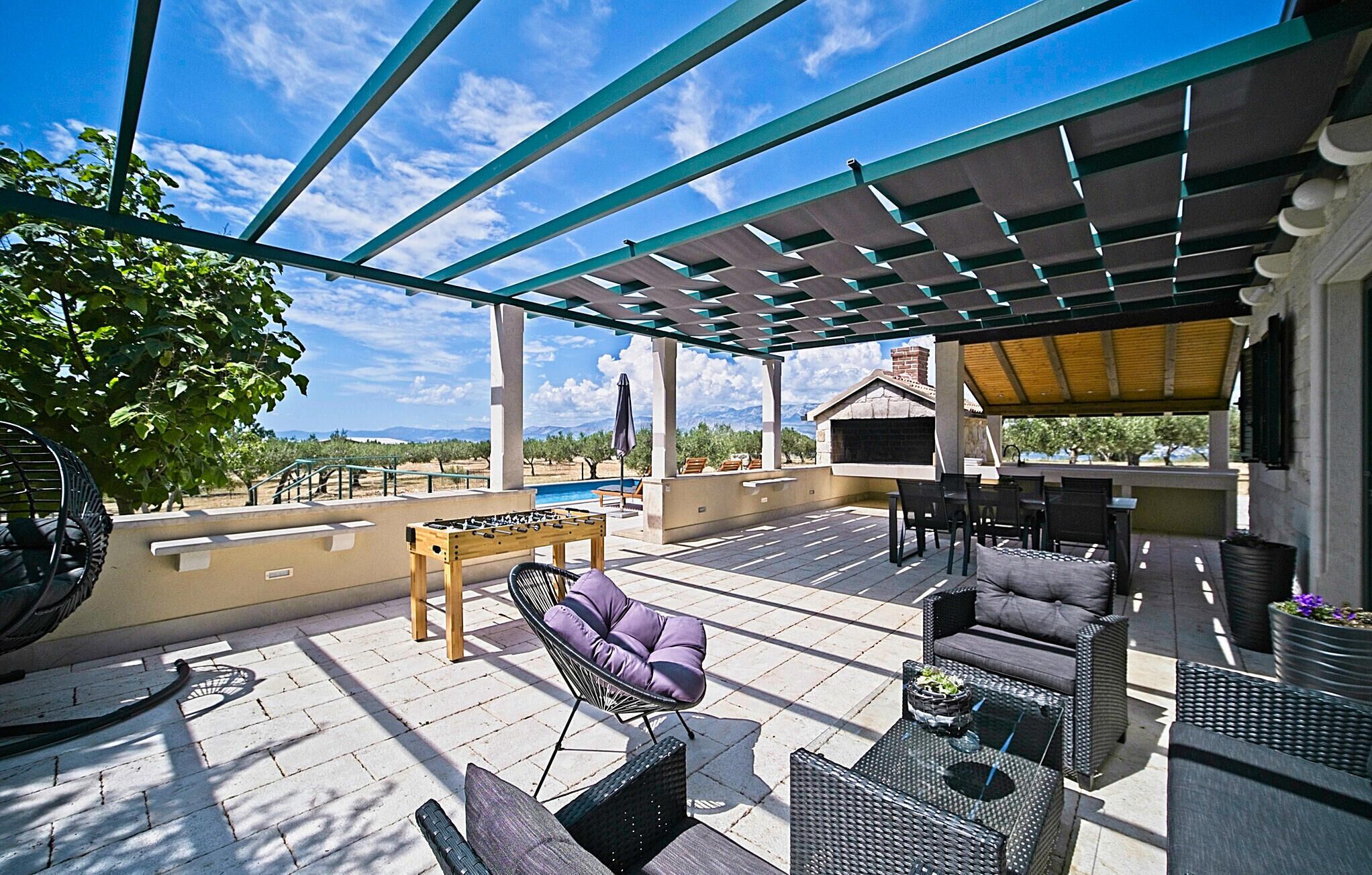 A covered outdoor patio with seating, a foosball table, and a pool in the background.