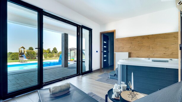 Ein modernes Zimmer mit einer großen Schiebeglastür, die zu einem Poolbereich, einem Whirlpool sowie einer Wasserrutsche und viel Grün führt.