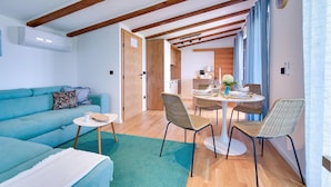 Ein modernes Wohnzimmer mit einem türkisfarbenen Sofa, einem Essbereich mit Korbstühlen und freiliegenden Holzbalken an der Decke.